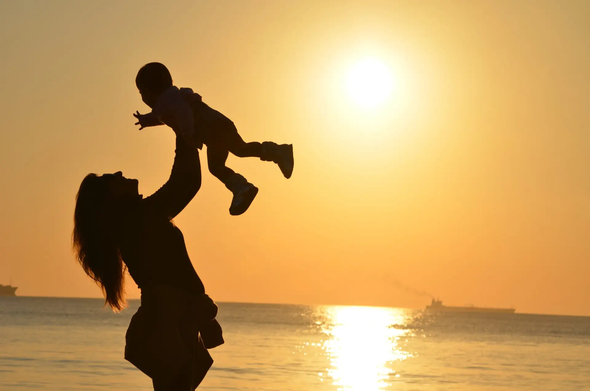 Woman holding child up on beach with sun in background